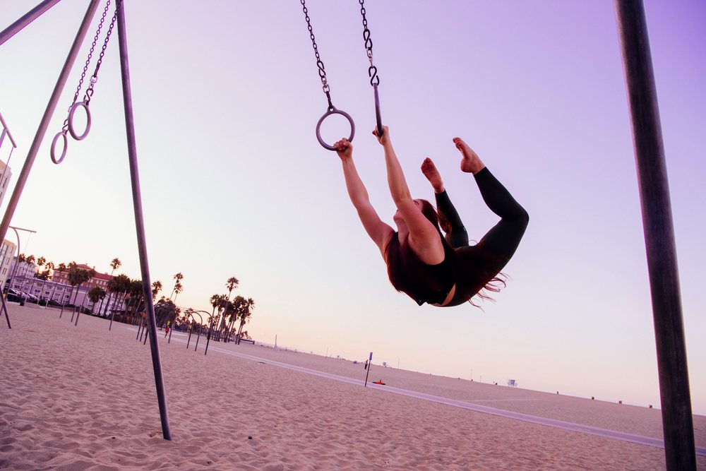 Woman hanging from gymnastics rings on a California beach at sunset, performing an exercise raising her legs outdoors, USA.