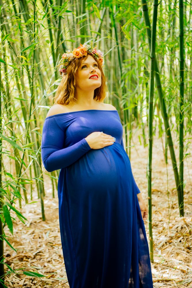 Pregnant woman with floral crown wearing a blue off-shoulder dress stands in a bamboo forest outdoors in natural light, pregnancy.