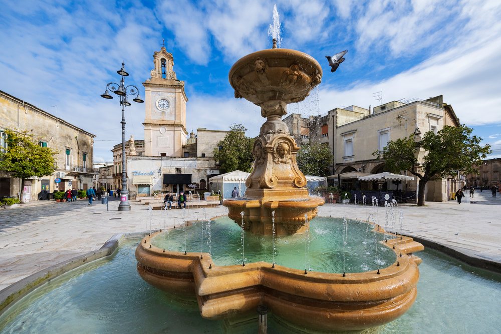 Fountain with water jets and a bird flying near the clock tower in Piazza Umberto, Francavilla Fontana, Puglia, Italy.