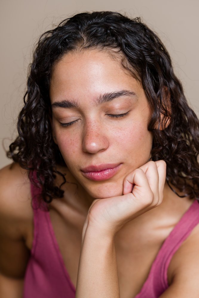 Portrait of a relaxed woman with closed eyes, curly hair, and freckles resting her hand on her chin against a plain background, beauty shoot, Spain.