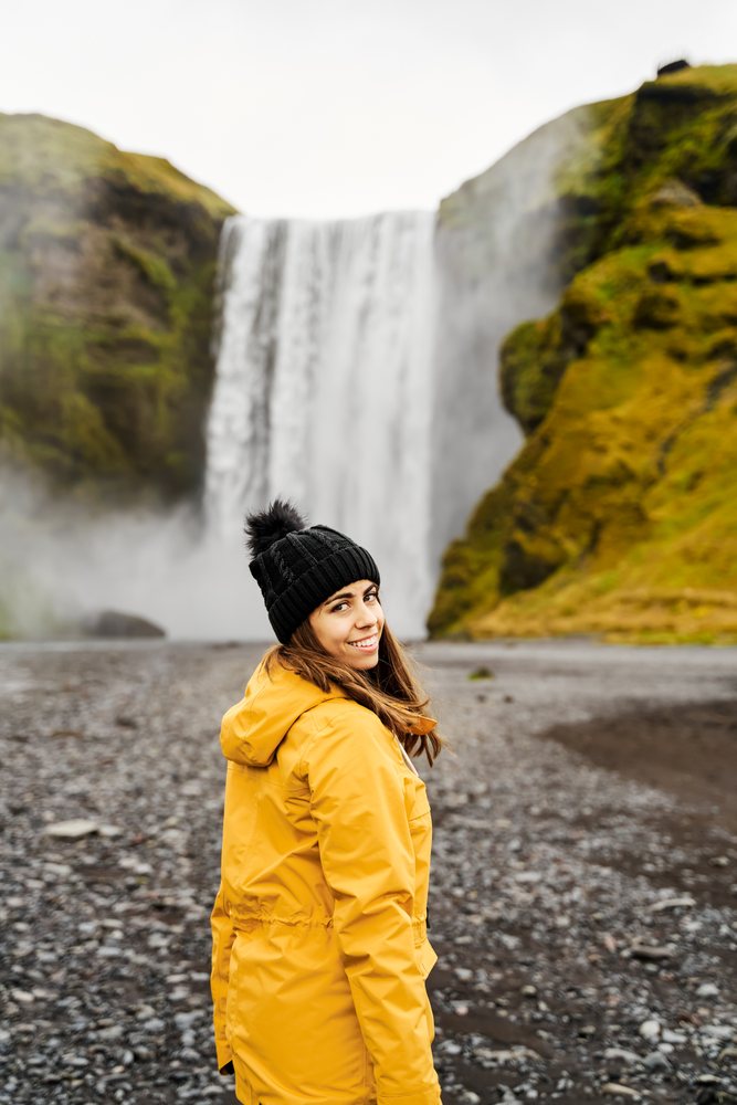 A woman wearing a yellow jacket and black beanie stands on rocky ground looking back at a waterfall on a misty day, Iceland.