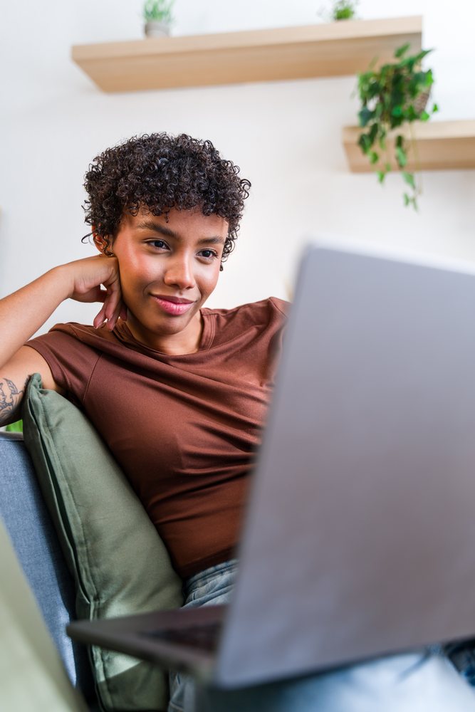 Woman with curly hair sitting on a couch using a laptop in a modern indoor setting with plants on wall shelves, Spain.