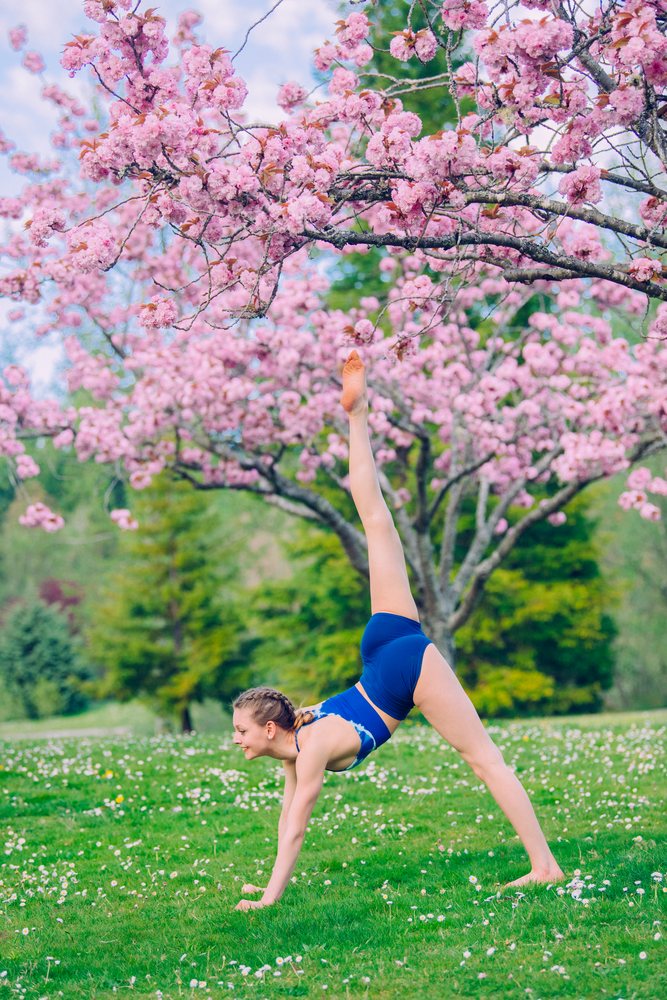 Woman performing a yoga pose with one leg raised near a flowering cherry tree in a grassy outdoor park, USA.