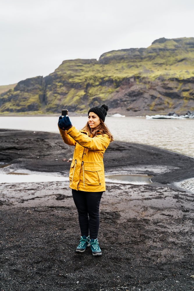 A white woman takes a selfie on volcanic black sand by a lake