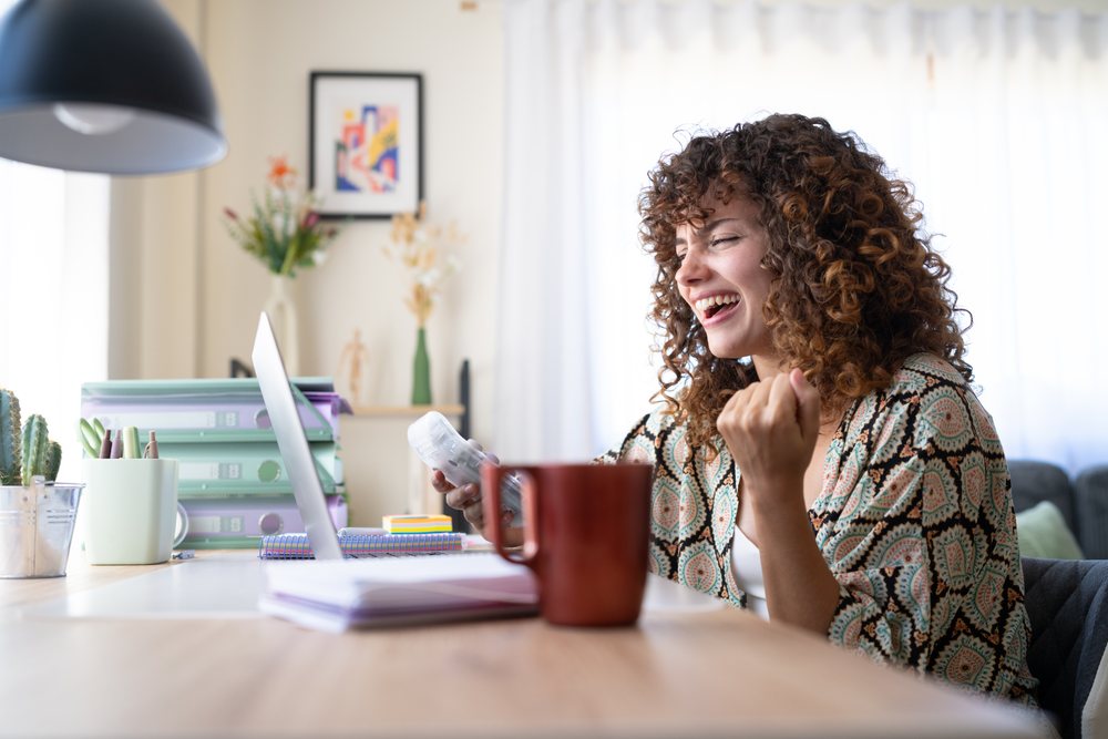 Woman with curly hair laughing and working at a wooden desk with a laptop and office supplies in a well-lit home office, Spain.