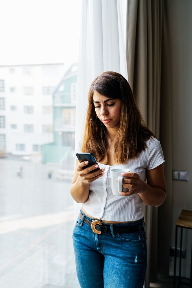 Woman using smartphone and holding cup indoors near large window