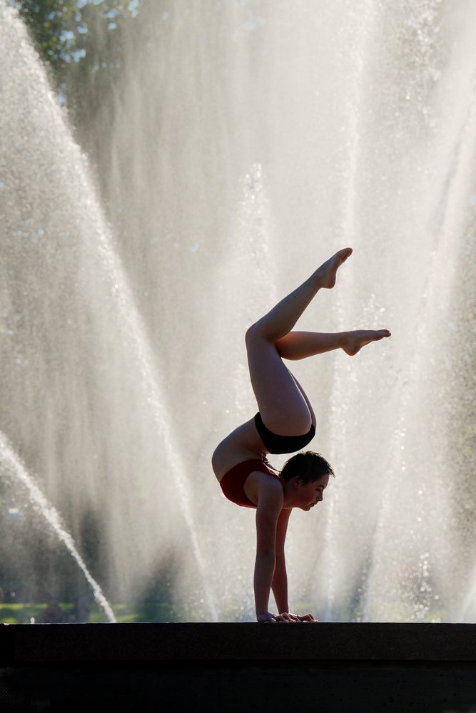 Young woman performs a handstand yoga pose outdoors by a large water fountain in the USA, silhouetted by sunlight.