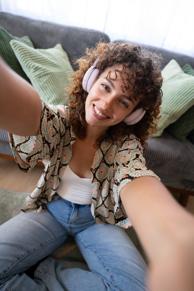 Woman with curly hair wearing headphones smiles while seated on a couch with green pillows during daytime, home, girl, Spain.