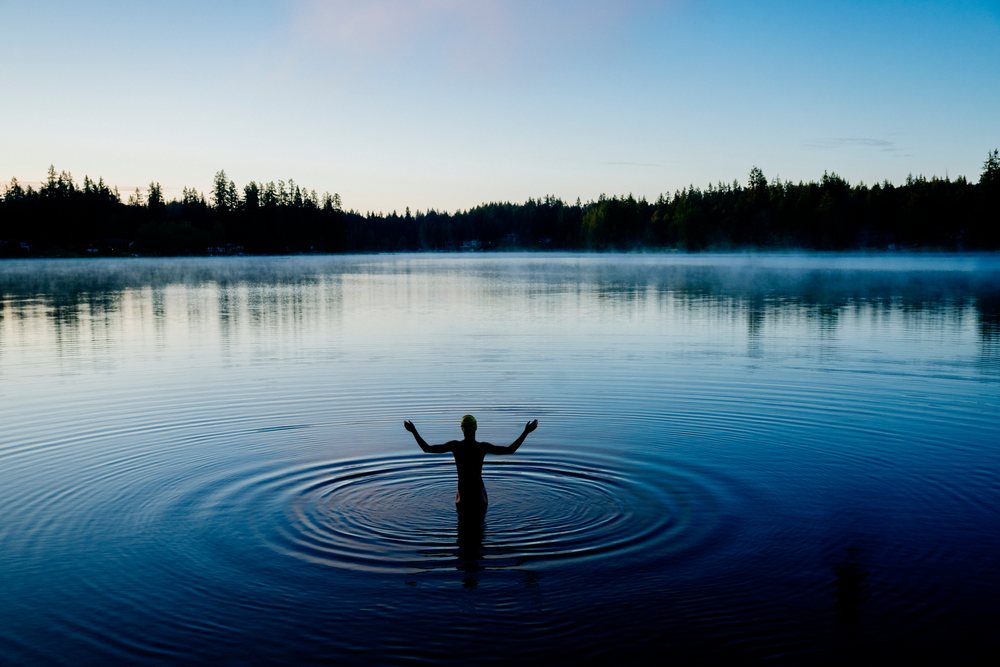 Person standing waist-deep in calm lake water surrounded by a misty tree-lined forest in the USA during early morning.