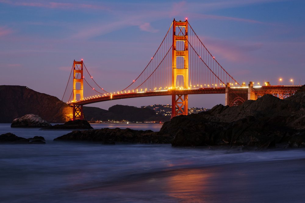Golden Gate Bridge illuminated at Dusk over the ocean with rocks, landscape, cityscape, city life, San Francisco, California, USA.