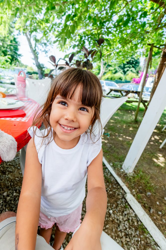 Smiling girl wearing pink shorts and a white sleeveless shirt enjoys a sunny day outdoors among green trees and picnic tables on gravel ground, kids, Spain.