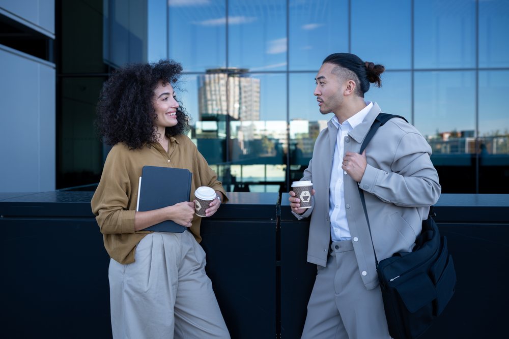 Two people holding coffee cups engage in conversation outdoors near a modern office building with glass reflections, Madrid, Spain.