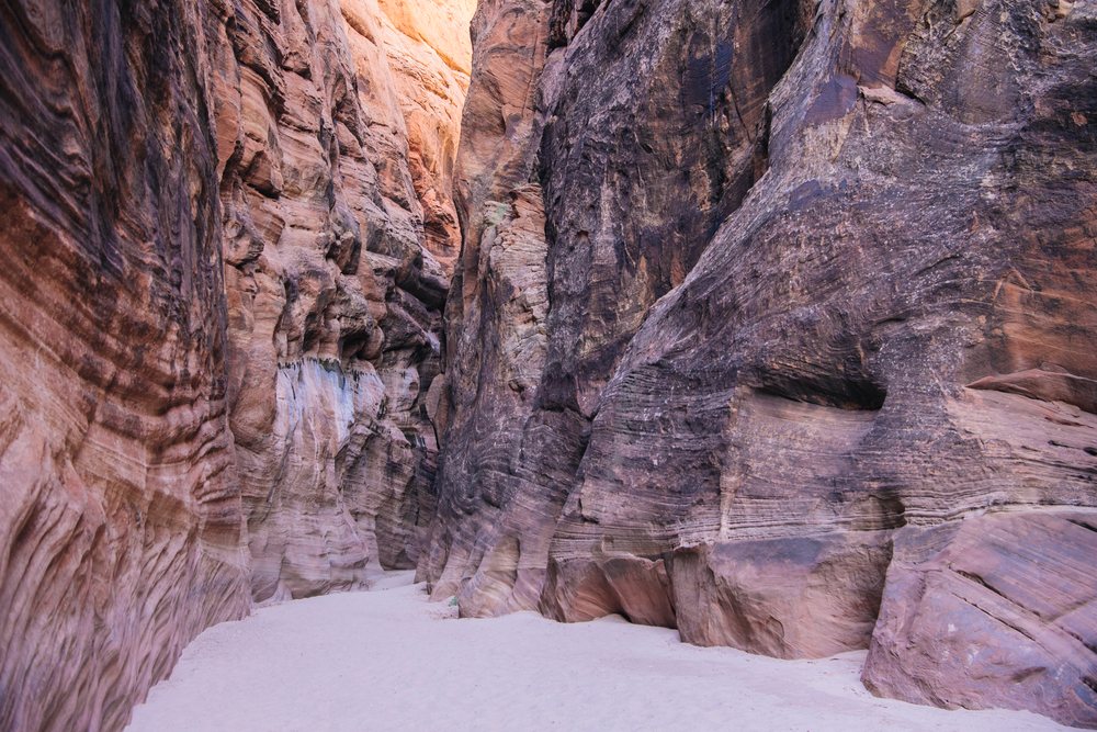 Buckskin Gulch features a narrow sandy pathway with towering sandstone rock walls displaying erosion patterns and sunlight highlights, slot canyon, Utah, USA.