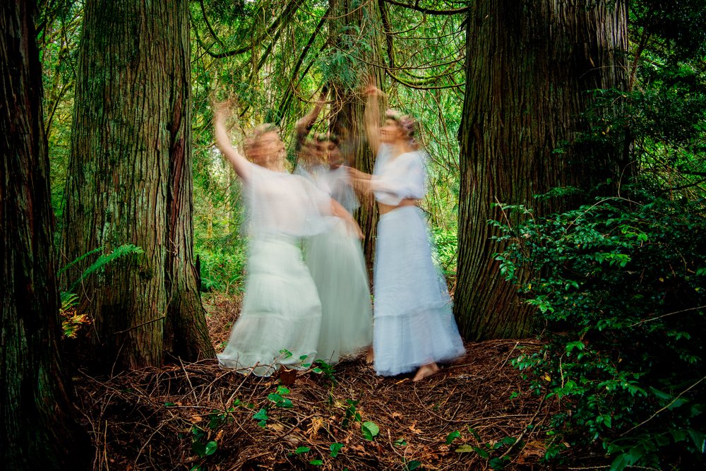 Three women dancing barefoot with motion blur in a green forest with tall trees, muses, USA.