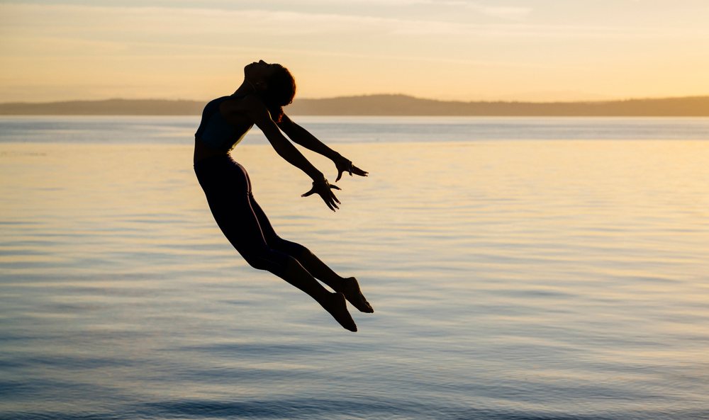 Woman in fitness attire gracefully leaps above a bench by a lakeshore at sunset, yoga, acrobatics, USA.