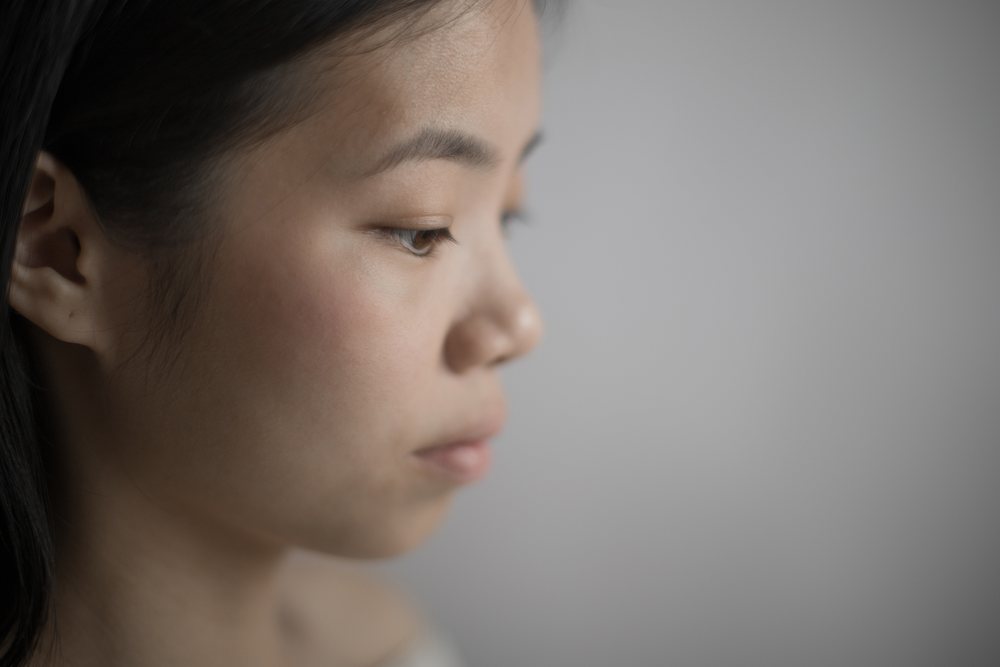 Profile portrait of an Asian woman with black hair and a contemplative, emotional expression against a neutral background.