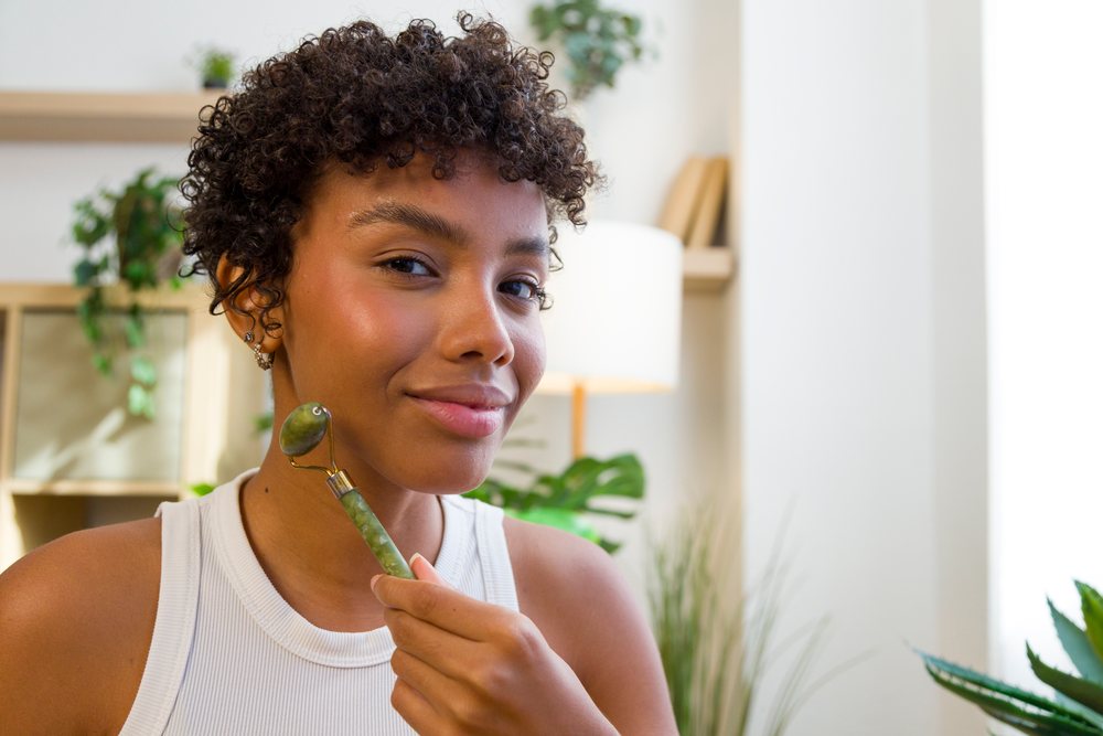 Young woman with curly hair uses a jade roller for facial massage in a home interior with indoor plants and natural light.