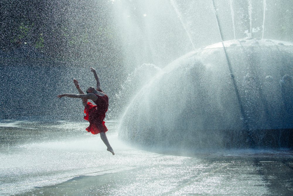 Female dancer in a red dress leaps gracefully through water spray from a large outdoor fountain in a USA park setting under sunlight.