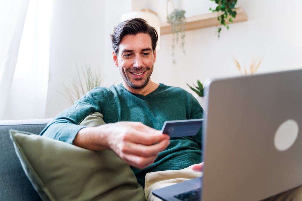 Man shopping online using a credit card with his laptop while sitting on a sofa at home, Spain.