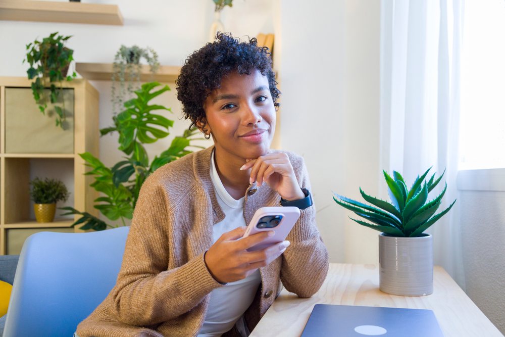 Young woman wearing brown cardigan holding a smartphone while seated at a desk with a laptop and potted succulent in a home living room.