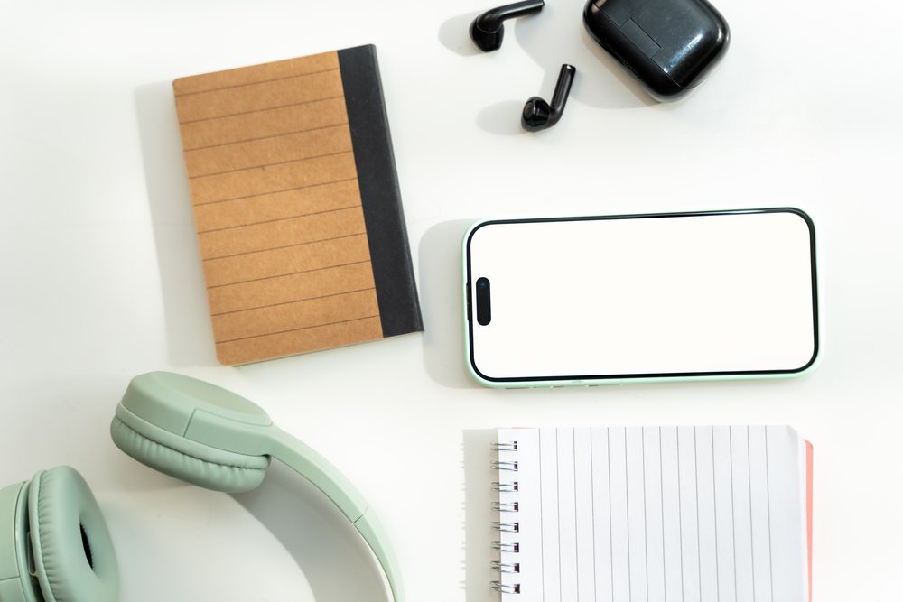 Smartphone with blank screen beside green headphones, black wireless earbuds, and brown and white notebooks on white desk, mock up, white screen, Spain.