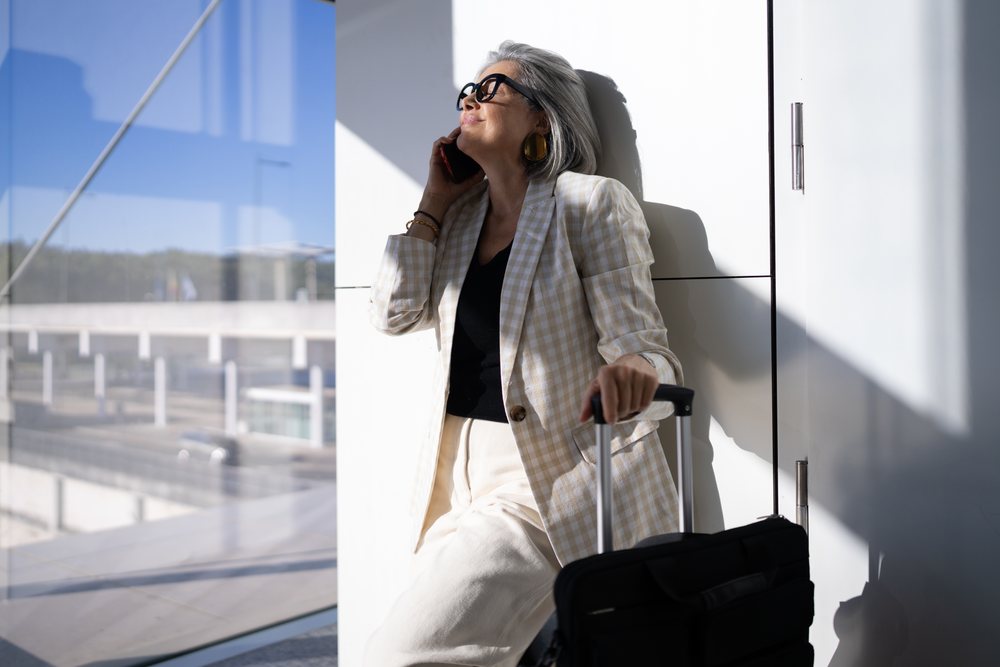 Woman with gray hair and glasses pulling a suitcase and talking on phone inside an airport terminal, businesswoman, traveler, Spain, Madrid.