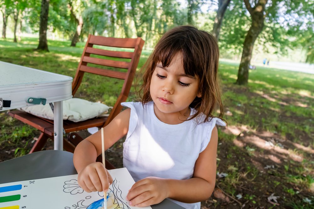 Child painting with a brush on paper at a table in a sunny park in Spain under trees, kids.