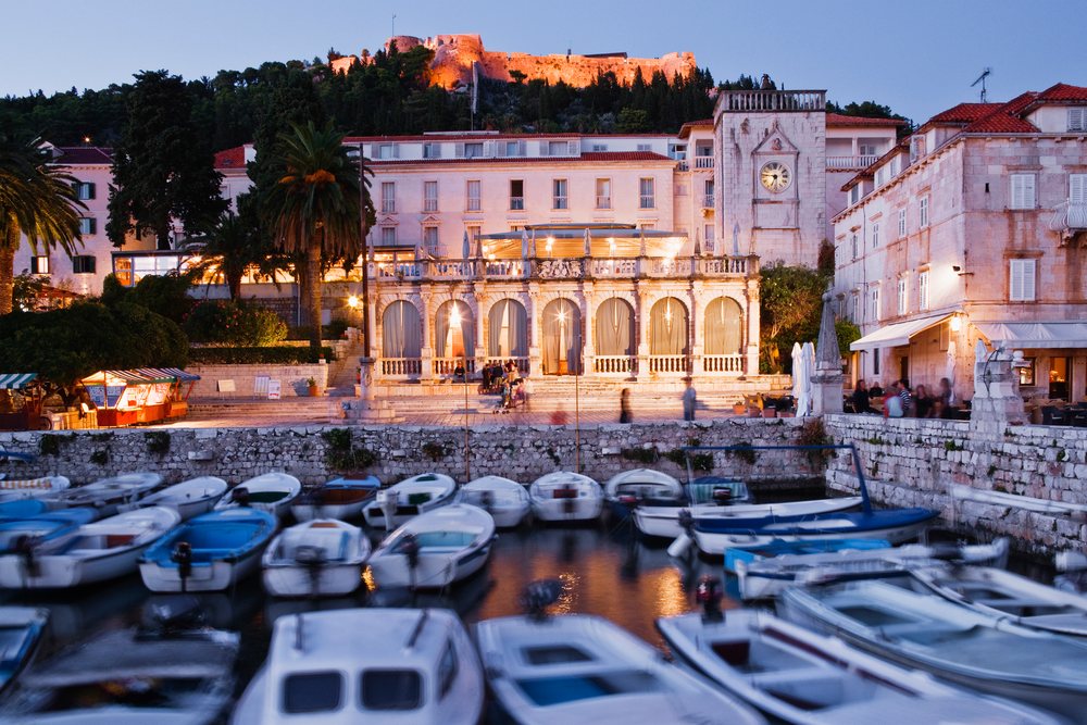 Multiple small boats dock in the calm harbour with illuminated historic buildings, a clock tower, and a hilltop fortress under an evening sky, Hvar Town, Croatia.