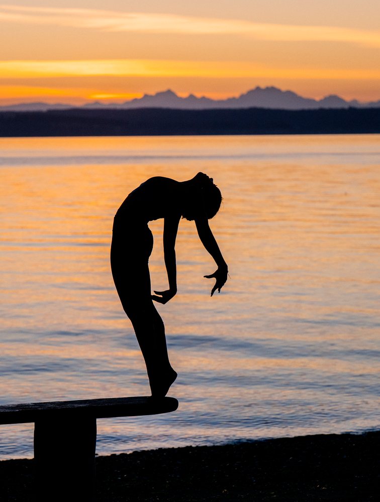 Silhouette of a woman performing a yoga backbend on a bench by the water at sunset, USA.