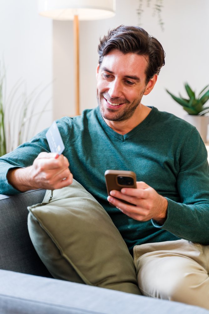 Smiling man in green sweater sitting on gray couch using smartphone and holding a credit card at home with green pillow and indoor plant visible, Spain.