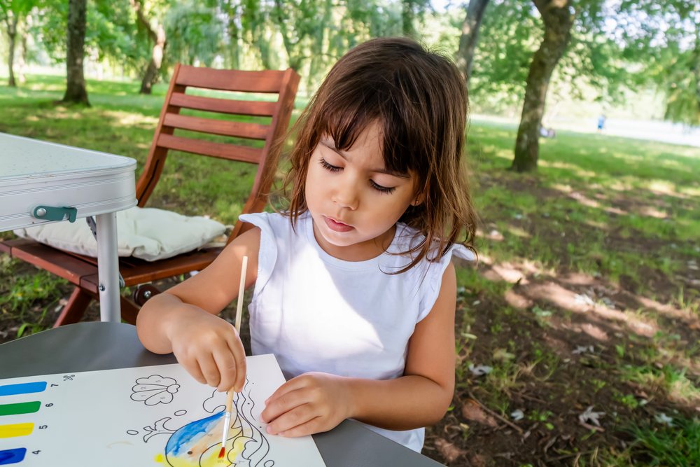 Child painting with watercolors on paper outdoors in a shaded park area with wooden chair and table nearby, kids, Spain.