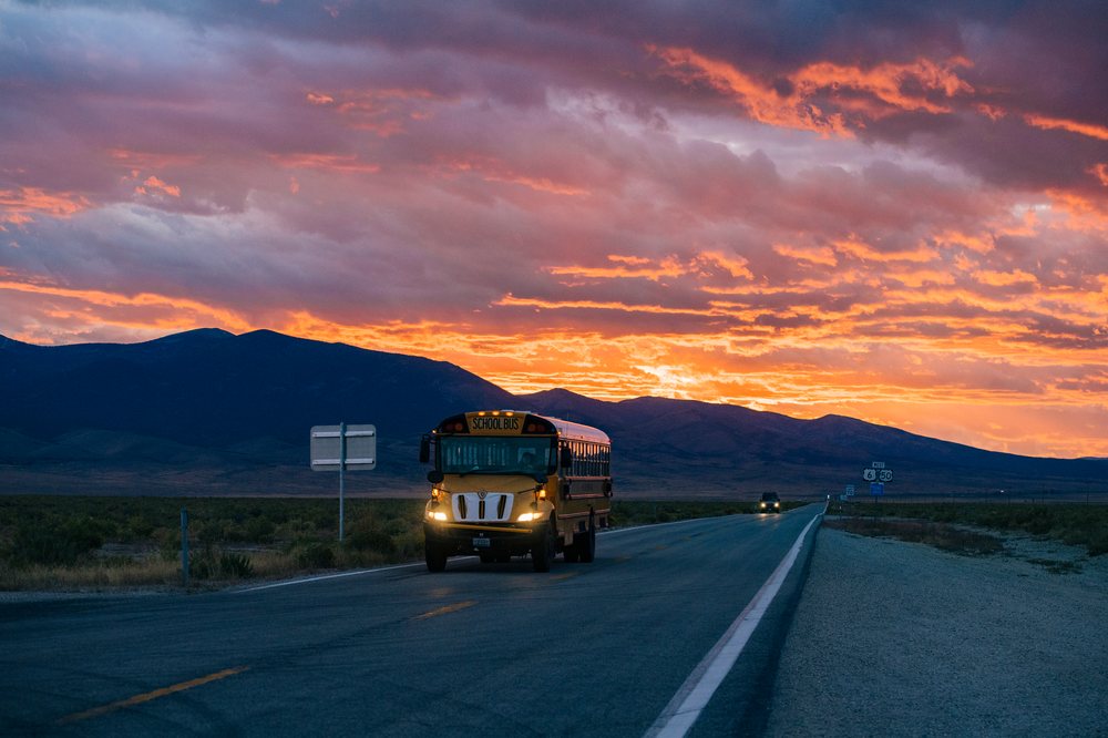 Yellow school bus driving on a paved highway at sunset with mountains in the background, Nevada, USA.