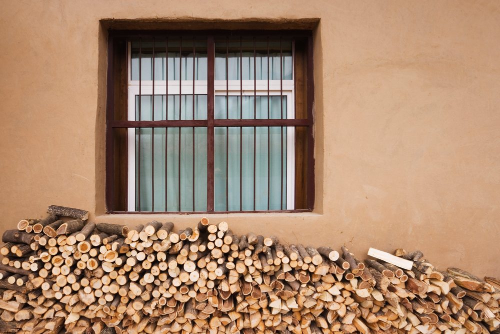 Stack of cut firewood logs piled below a barred window on a brown plaster wall, Shaanxi Province, China.