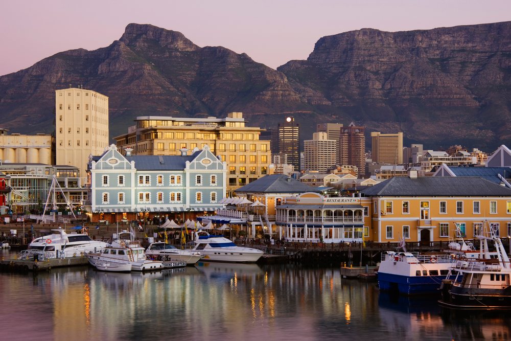 Boats docked at a waterfront harbor with buildings and Table Mountain in the background at sunset, South Africa.