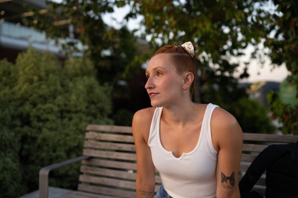 A young woman with a cat tattoo on her arm sits on a wooden bench outdoors in a park during sunset, urban, Santiago De Compostela, Spain.