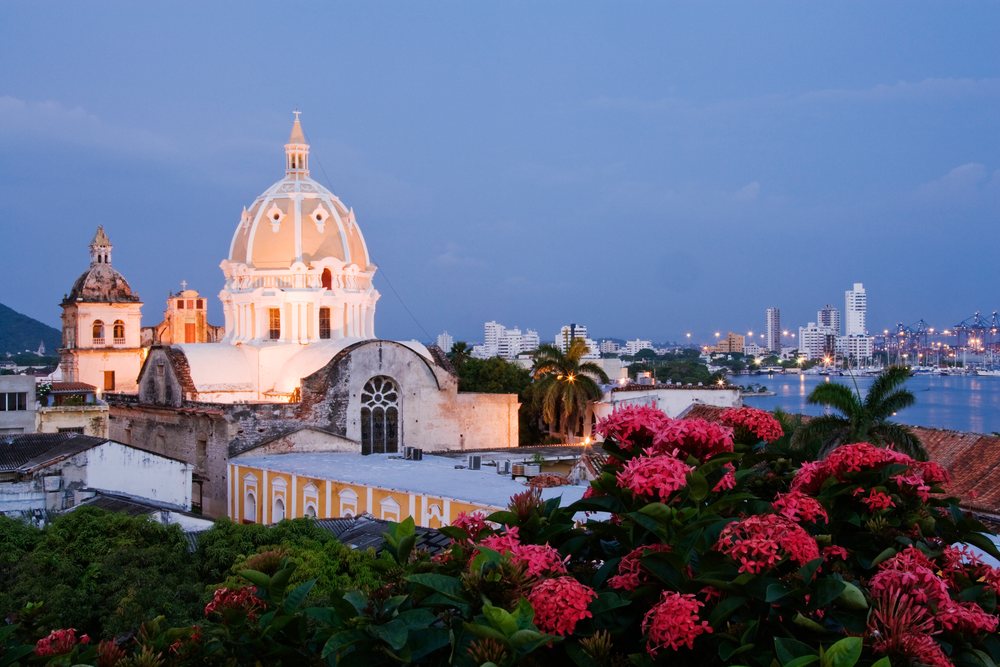 Historic Iglesia de San Pedro Claver with illuminated dome and city skyline at dusk, with red flowers foreground, Cartagena, Colombia.