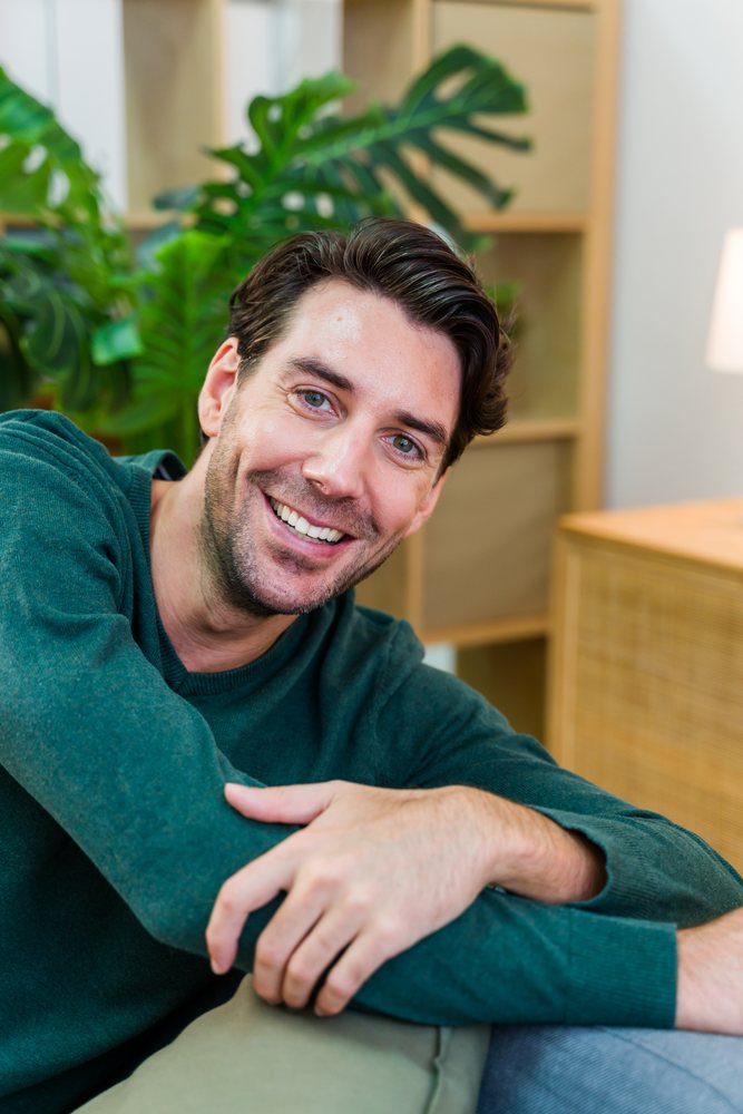 Man in a green sweater smiling indoors while seated on a couch near houseplants and wooden furniture, Spain.