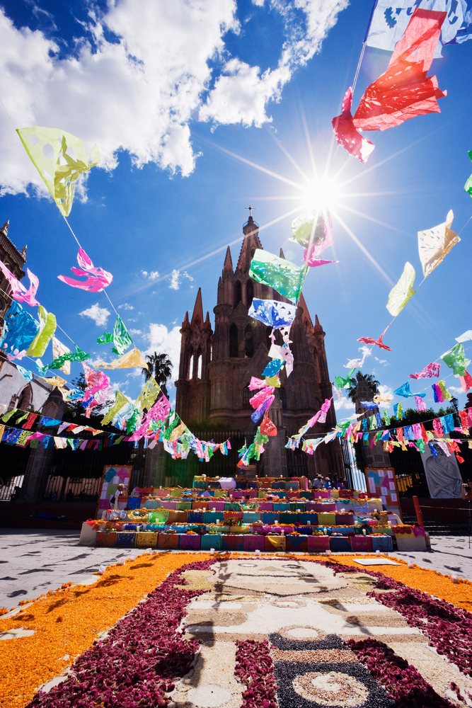 Day of the Dead altar decorated with colorful papel picado and marigold petals in front of San Miguel de Allende cathedral under bright sunlight, Mexico.