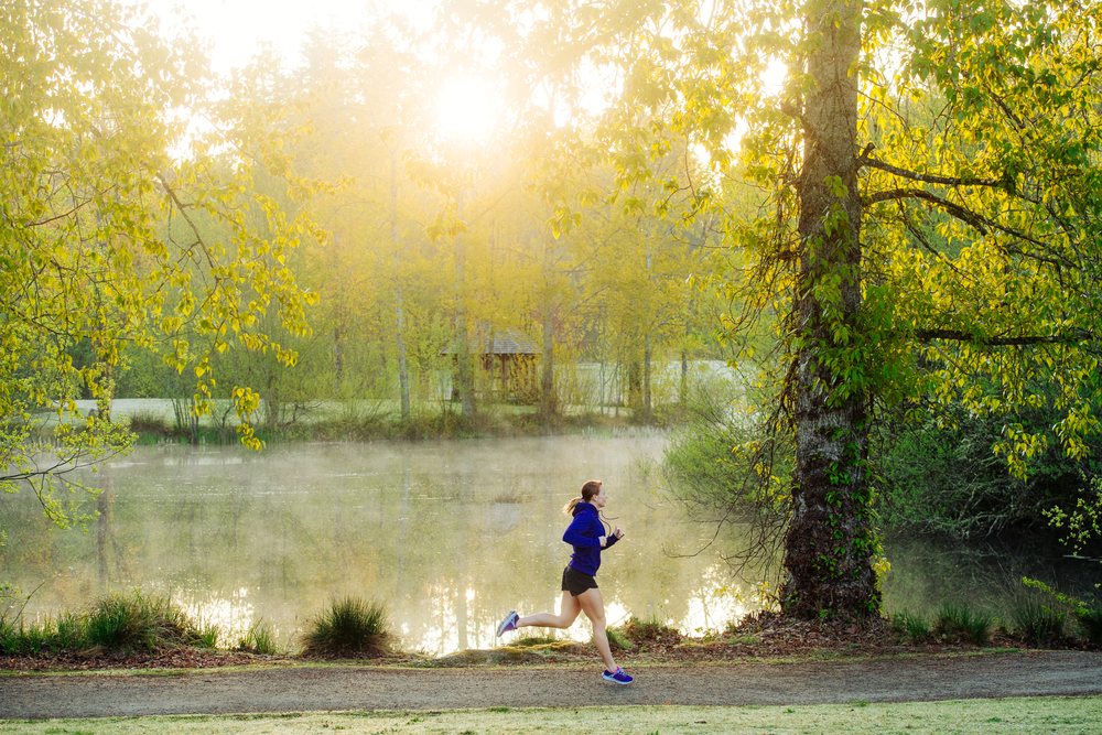 Woman running on a trail in a park by a lake with morning sunlight and mist, Washington, Bainbridge Island, USA.