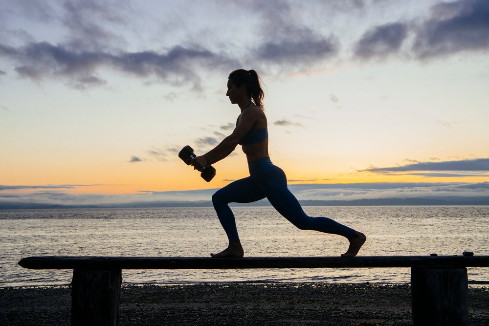 Woman exercising barefoot on a wooden bench by the water at sunset, WA, USA.