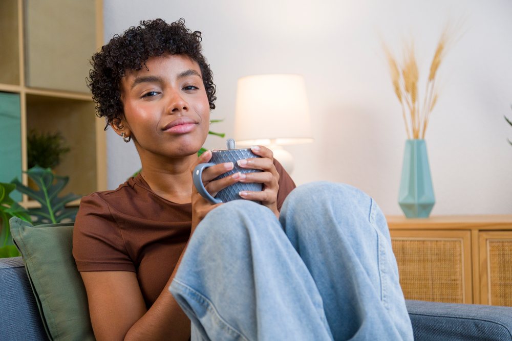 Young woman sitting on a sofa holding a mug comfortably in a cozy indoor living room with lamp, green pillow, and decorative plants.