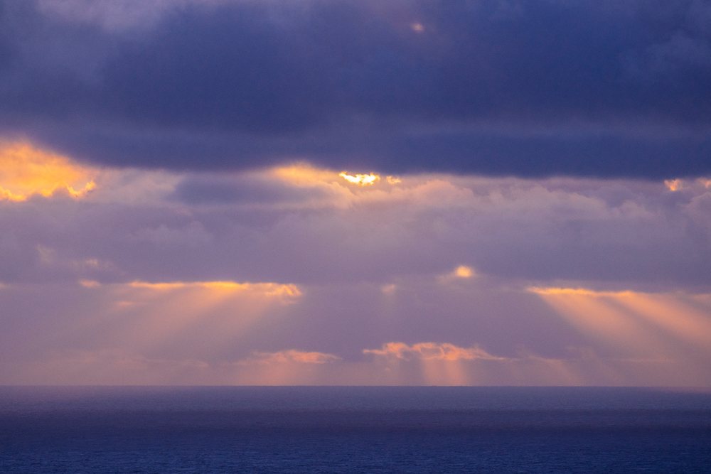 Sunlight streams through layered clouds over the ocean horizon during a calm sunrise, California, USA.