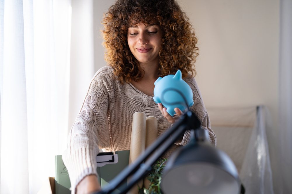 Smiling woman holding blue piggy bank while organizing items on desk in a well-lit indoor space, moving, Spain.