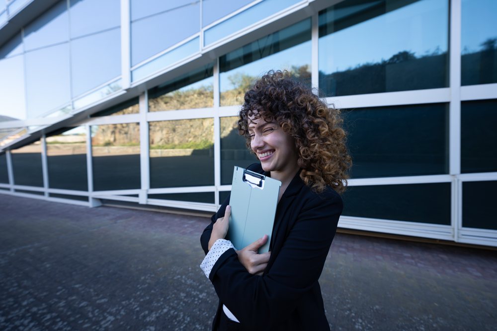 Businesswoman smiling and holding a clipboard outside a modern glass building, business trip, Spain, Santiago De Compostela.