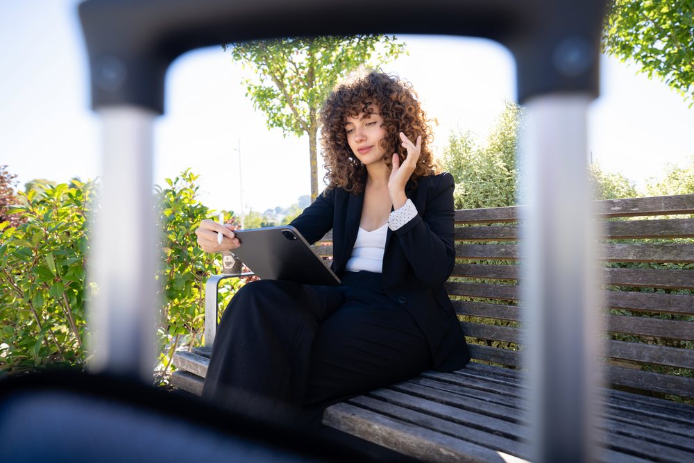 Woman in business attire sitting on wooden bench using tablet with digital pen during daytime, Spain.