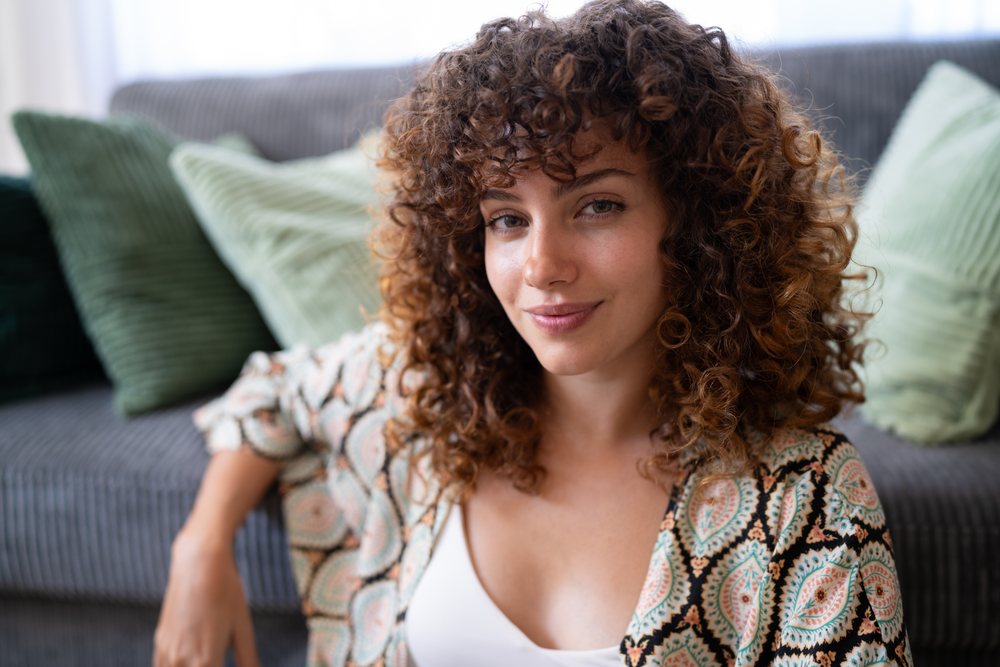 Woman with curly hair smiling while seated on a sofa with green cushions in an indoor home setting, Spain.