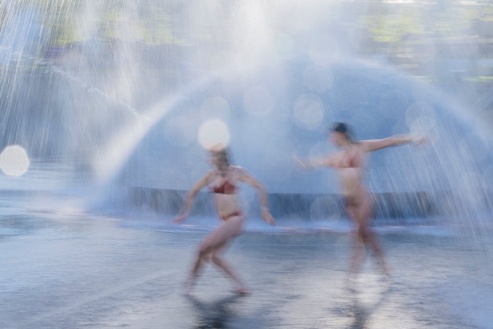 Two women in bikinis dancing and moving dynamically through water sprays at an outdoor water fountain, partner, acrobatics, yoga, USA.