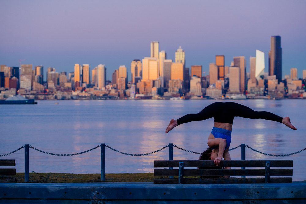 Woman performing a handstand yoga pose on a wooden bench by the Seattle waterfront at sunset with calm ter reflections, WA, USA.