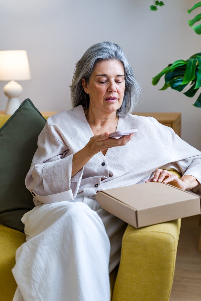 Woman with gray hair sits on a yellow couch speaking into a smartphone with a brown cardboard box on her lap indoors, Spain.