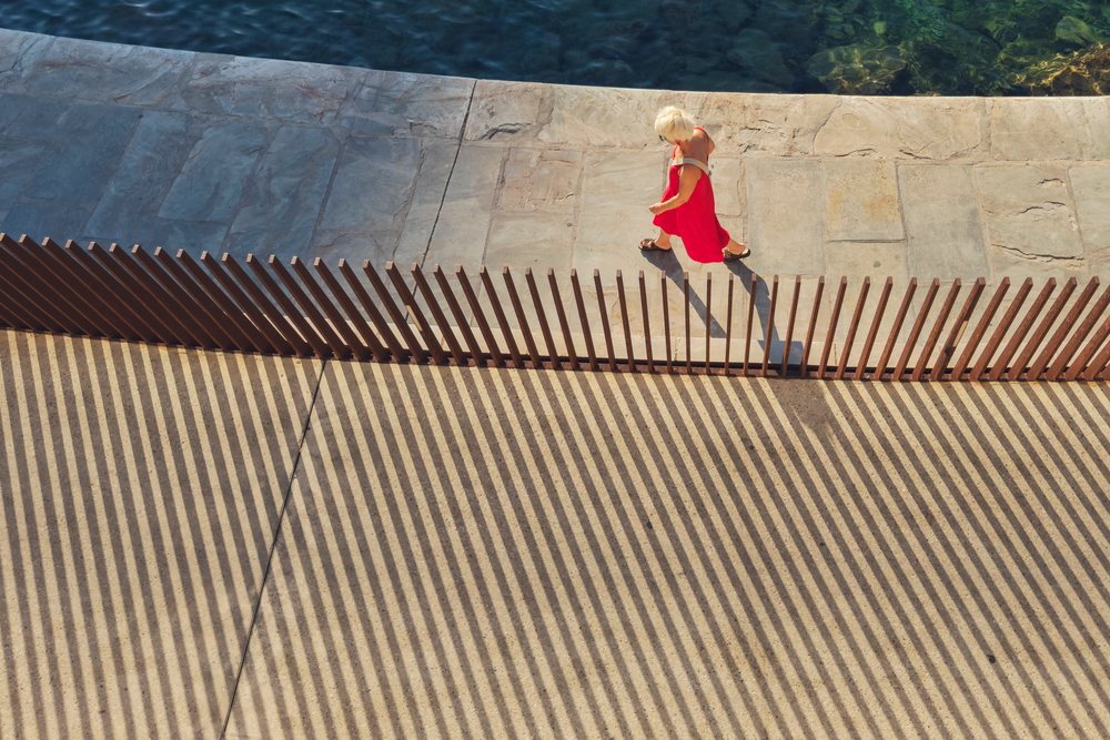 Woman in a red dress walks along a stone breakwater with wooden fence shadows in bright sunlight, Travel Photography, Bastia, Corsica, France.
